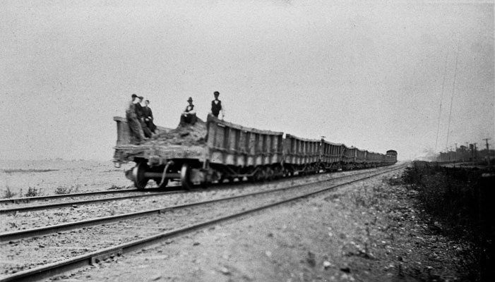 <p>Workers casually sit atop a pile of earth excavated from the canal work site. Hundreds of these trains would on a daily basis be transporting material to be used in lock construction, or the building of canal embankments or the breakwaters in the lakes. Loads of earth might also be deposited in waste dumps forming berms set back from the canal. Photo attribution: St. Catharines Museum, 2013-N</p>